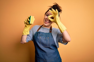 Young brunette cleaner woman wearing housekeeping gloves holding scourer scrub with happy face...