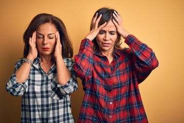 Middle age beautiful couple of sisters wearing casual shirt over isolated yellow background suffering from headache desperate and stressed because pain and migraine. Hands on head.