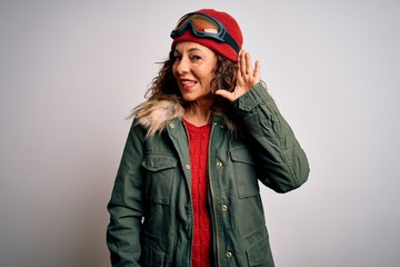 Middle age skier woman wearing snow sportswear and ski goggles over white background smiling with hand over ear listening an hearing to rumor or gossip. Deafness concept.