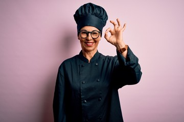 Middle age brunette chef woman wearing cooker uniform and hat over isolated pink background smiling positive doing ok sign with hand and fingers. Successful expression.