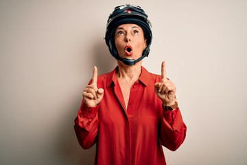 Middle age motorcyclist woman wearing motorcycle helmet over isolated white background amazed and surprised looking up and pointing with fingers and raised arms.