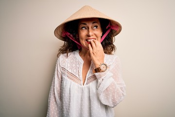 Middle age brunette woman wearing asian traditional conical hat over white background looking stressed and nervous with hands on mouth biting nails. Anxiety problem.