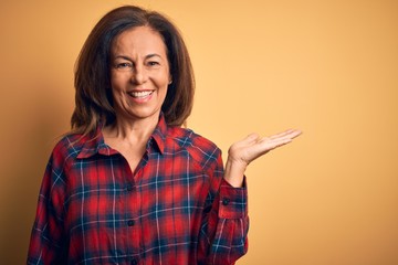 Middle age beautiful woman wearing casual shirt standing over isolated yellow background smiling cheerful presenting and pointing with palm of hand looking at the camera.