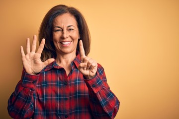 Middle age beautiful woman wearing casual shirt standing over isolated yellow background showing and pointing up with fingers number six while smiling confident and happy.