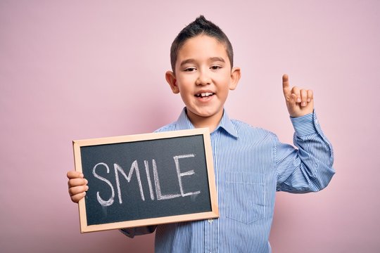 Young little boy kid showing blackboard with smile word as happy message over pink background surprised with an idea or question pointing finger with happy face, number one