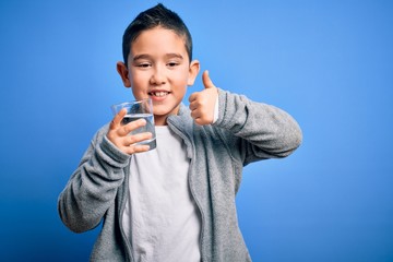 Adorable toddler smiling happy and confident. Standing with smile on face holding glass of healthy water to refreshment with thumb up doing ok sign over isolated blue background