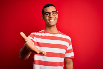 Young handsome african american man wearing casual striped t-shirt and glasses smiling cheerful offering palm hand giving assistance and acceptance.