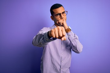 Handsome african american man wearing striped shirt and glasses over purple background Punching fist to fight, aggressive and angry attack, threat and violence