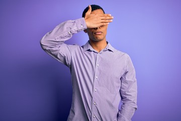 Handsome african american man wearing striped shirt and glasses over purple background covering eyes with hand, looking serious and sad. Sightless, hiding and rejection concept