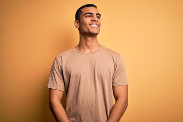 Young handsome african american man wearing casual t-shirt standing over yellow background looking away to side with smile on face, natural expression. Laughing confident.