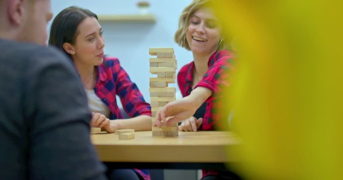 Happy group of friends is playing jenga, a pretty girl takes a bar from jenga and laughs. Girl carefully pulls the bar out of the jenga, she is happy. Company plays board games and having fun.