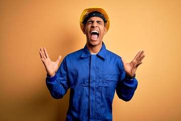 Young handsome african american worker man wearing blue uniform and security helmet celebrating mad...