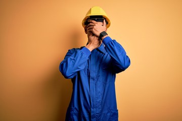 Young handsome african american worker man wearing blue uniform and security helmet Covering eyes and mouth with hands, surprised and shocked. Hiding emotion