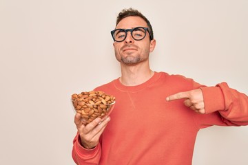 Handsome man with blue eyes holding bowl with healthy almonds snack over white background with surprise face pointing finger to himself