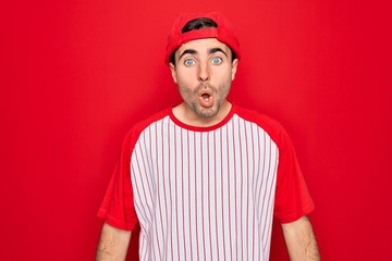 Young handsome sporty man with blue eyes wearing striped baseball t-shirt and cap afraid and shocked with surprise expression, fear and excited face.