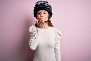 Young beautiful motorcyclist woman with blue eyes wearing moto helmet over pink background looking at the camera blowing a kiss with hand on air being lovely and sexy. Love expression.