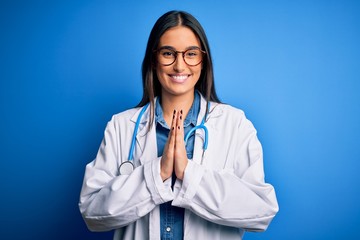 Young beautiful doctor woman wearing stethoscope and glasses over blue background praying with hands together asking for forgiveness smiling confident.