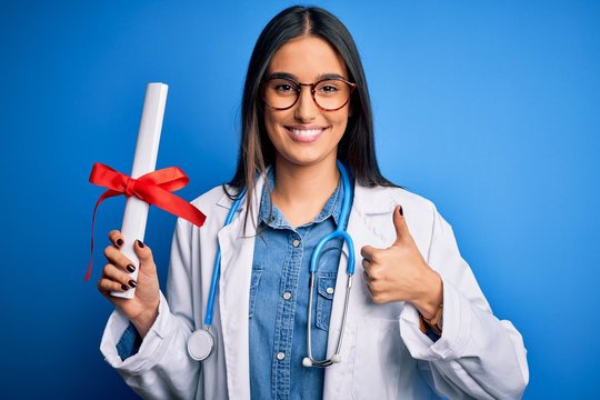 Young beautiful brunette doctor woman wearing glasses and coat holding diploma degree happy with big smile doing ok sign, thumb up with fingers, excellent sign