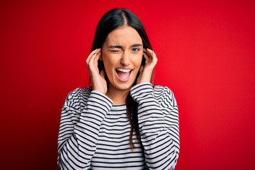 Young beautiful brunette woman wearing casual striped t-shirt over red background covering ears with fingers with annoyed expression for the noise of loud music. Deaf concept.