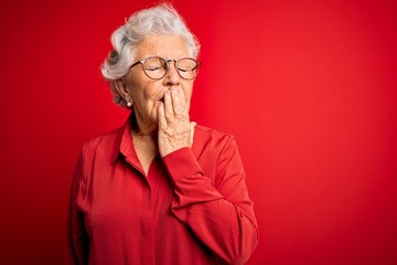 Senior beautiful grey-haired woman wearing casual shirt and glasses over red background bored yawning tired covering mouth with hand. Restless and sleepiness.