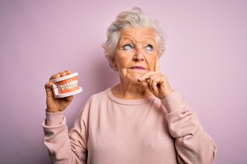 Senior beautiful grey-haired woman holding plastic denture teeth over pink background serious face thinking about question, very confused idea