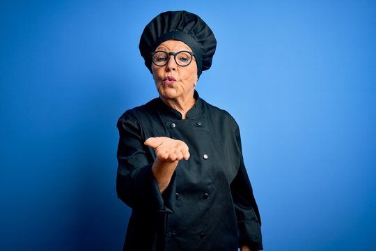 Senior Beautiful Grey-haired Chef Woman Wearing Cooker Uniform And Hat Over Blue Background Looking At The Camera Blowing A Kiss With Hand On Air Being Lovely And Sexy. Love Expression.