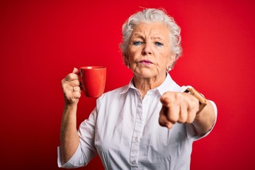 Senior beautiful woman drinking mug of coffee standing over isolated red background pointing with...