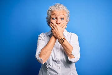 Senior beautiful woman wearing elegant shirt standing over isolated blue background shocked covering mouth with hands for mistake. Secret concept.
