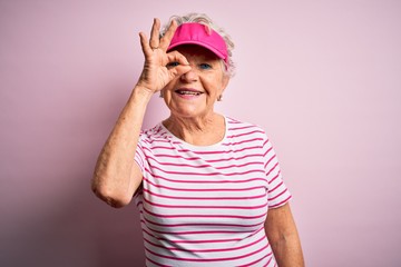 Senior beautiful sporty woman wearing sport cap standing over isolated pink background doing ok gesture with hand smiling, eye looking through fingers with happy face.