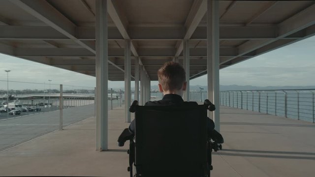 Middle shot of following the child in electric wheelchair enjoying the time outdoor. Handicapped boy moving along the waterfront