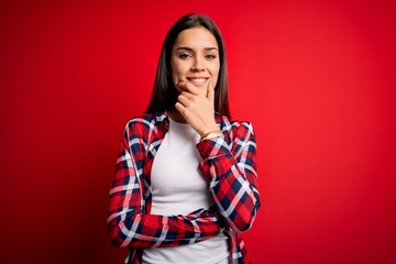 Young beautiful brunette woman wearing casual shirt standing over isolated red background looking confident at the camera smiling with crossed arms and hand raised on chin. Thinking positive.