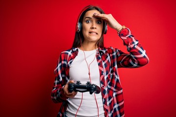 Young beautiful brunette gamer woman playing video game using joystick and headphones stressed with hand on head, shocked with shame and surprise face, angry and frustrated. Fear and upset