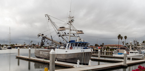 Obraz premium Harbor docks in Ventura sit over calm water with squid fishing boat moored under overcast cloudy sky.
