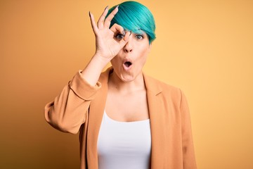 Young beautiful woman with blue fashion hair wearing casual jacket over yellow background doing ok gesture shocked with surprised face, eye looking through fingers. Unbelieving expression.