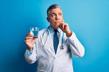 Middle age handsome grey-haired doctor man drinking glass of water over blue background serious face thinking about question, very confused idea