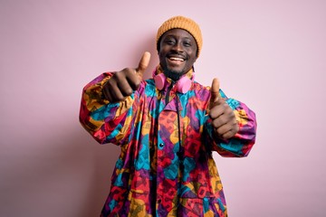 Young handsome african american man wearing colorful coat and cap over pink background approving doing positive gesture with hand, thumbs up smiling and happy for success. Winner gesture.