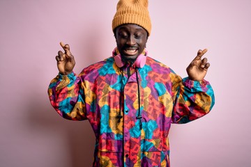 Obraz premium Young handsome african american man wearing colorful coat and cap over pink background gesturing finger crossed smiling with hope and eyes closed. Luck and superstitious concept.
