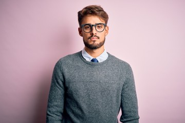 Young handsome man with beard wearing glasses and sweater standing over pink background Relaxed with serious expression on face. Simple and natural looking at the camera.