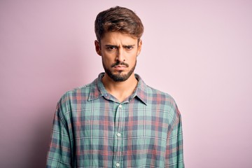 Young handsome man with beard wearing casual shirt standing over pink background skeptic and...
