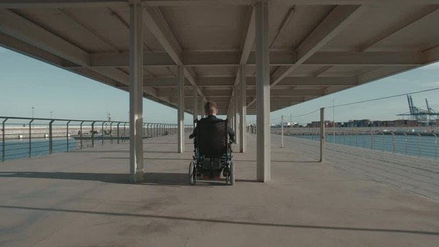 Wide back shot of a disabled child in electric wheelchair riding along the waterfront. He wants to breathe fresh air as other people