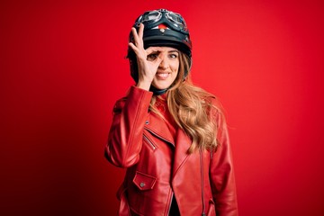 Young beautiful brunette motrocyclist woman wearing moto helmet over red background doing ok gesture with hand smiling, eye looking through fingers with happy face.