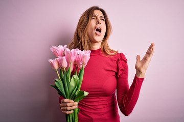 Young beautiful brunette woman holding bouquet of pink tulips over isolated background crazy and mad shouting and yelling with aggressive expression and arms raised. Frustration concept.