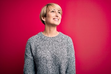 Young blonde woman with modern short hair wearing casual sweater over pink background looking away to side with smile on face, natural expression. Laughing confident.