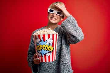 Young blonde woman with short hair wearing 3d movie glasses and eating popcorn at the cinema stressed with hand on head, shocked with shame and surprise face, angry and frustrated. Fear and upset for 
