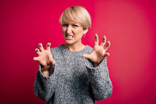 Young blonde woman with modern short hair wearing casual sweater over pink background smiling funny doing claw gesture as cat, aggressive and sexy expression