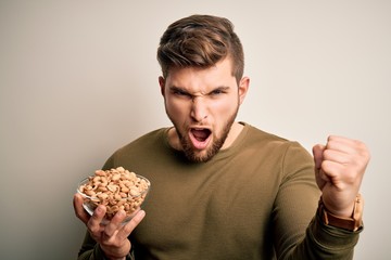 Young blond man with beard and blue eyes holding bowl with healthy salty peanuts annoyed and frustrated shouting with anger, crazy and yelling with raised hand, anger concept