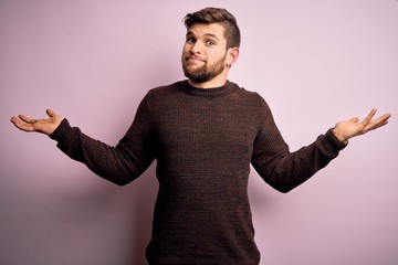 Young blond man with beard and blue eyes wearing casual sweater over pink background clueless and...