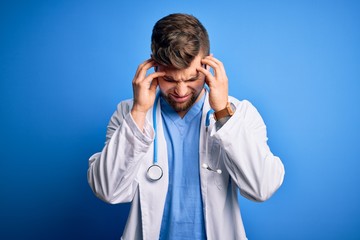 Young blond doctor man with beard and blue eyes wearing white coat and stethoscope with hand on head for pain in head because stress. Suffering migraine.