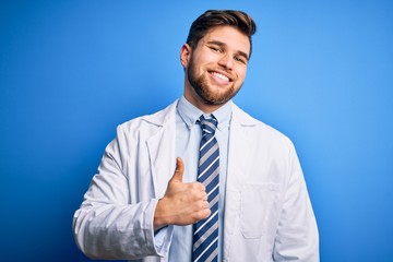 Young blond therapist man with beard and blue eyes wearing coat and tie over background doing happy thumbs up gesture with hand. Approving expression looking at the camera showing success.