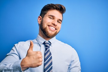 Young blond businessman with beard and blue eyes wearing elegant shirt and tie standing doing happy thumbs up gesture with hand. Approving expression looking at the camera showing success.